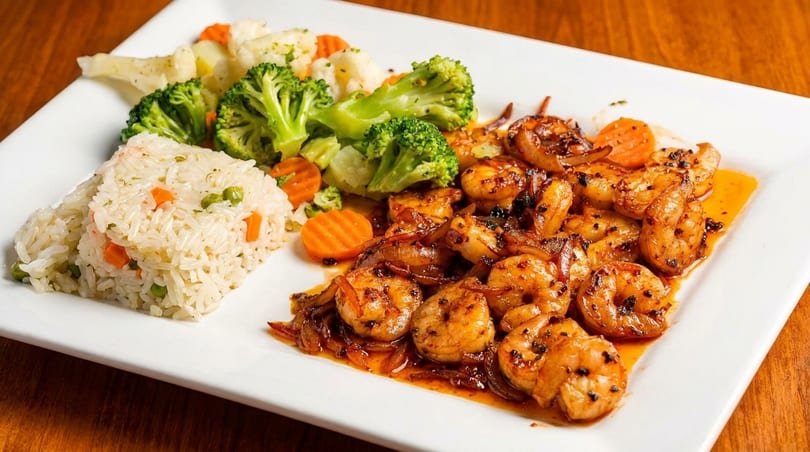 A white plate with seasoned shrimp and onions, rice pilaf, and steamed broccoli, cauliflower, and carrots.