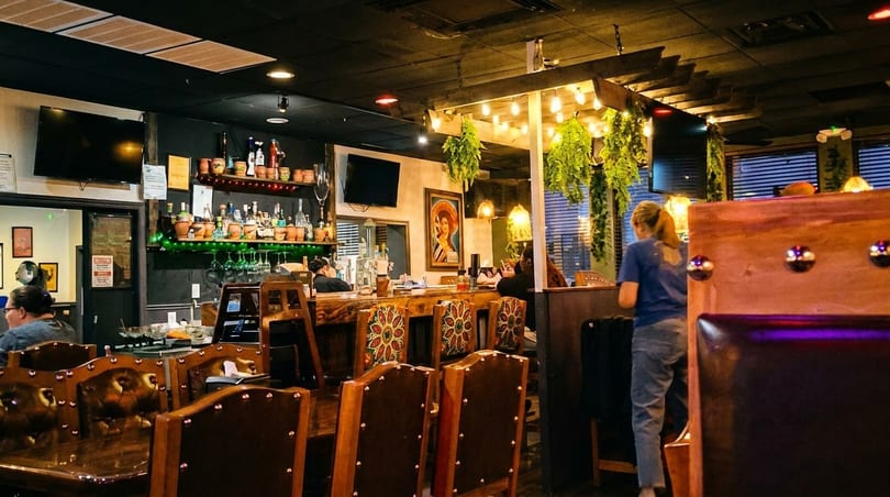A restaurant interior with a bar area, patrons, and decorative plants hanging from the ceiling.